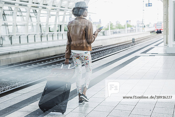Rückansicht der jungen Frau mit Smartphone und Gepäck am Bahnsteig