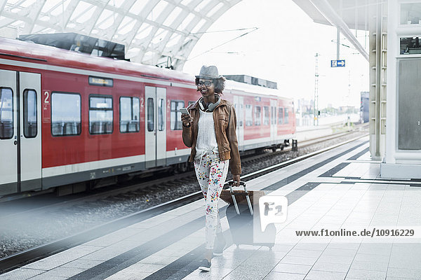 Junge Frau mit Trolleytasche und Smartphone am Bahnsteig