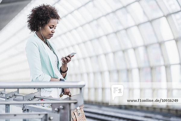 Junge Frau mit Kopfhörern schaut auf ihr Smartphone  während sie auf dem Bahnsteig wartet.