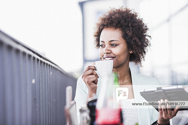 Lächelnde junge Frau mit Tasse Kaffee und digitalem Tablett in einem Straßencafé