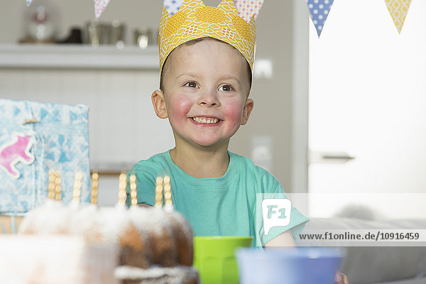 Glücklicher Junge mit Papierkrone am Geburtstagstisch sitzend