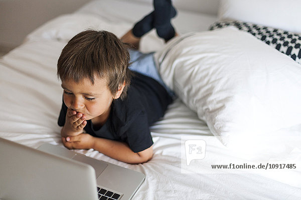 Smiling little boy lying on bed using laptop