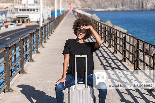 Portrait of smiling woman sitting on her rolling suitcase on a jetty