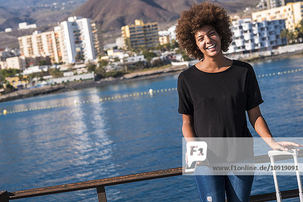 Spain  Tenerife  portrait of laughing young woman standing on deck with smartphone and suitcase