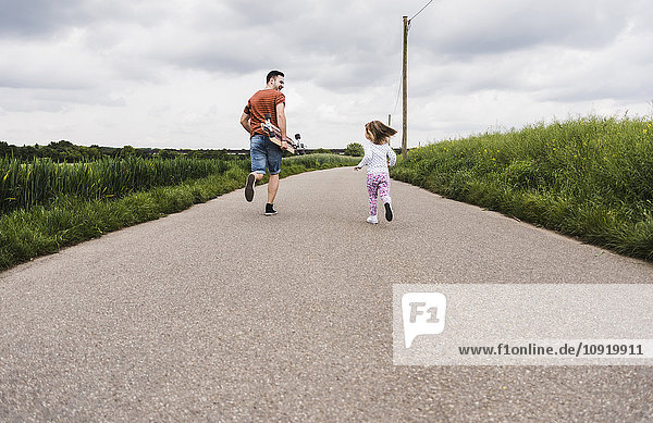 Father with skateboard and daughter running on country lane