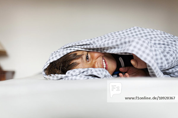 Lächelnder kleiner Junge  der unter der Decke auf seinem Bett liegt.