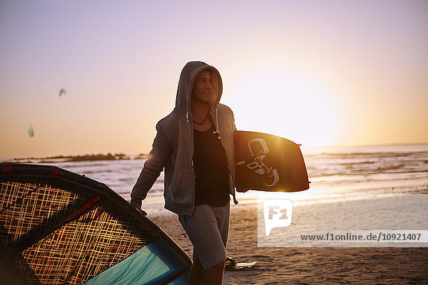 Mann in Kapuze mit Kiteboardausrüstung am Strand bei Sonnenuntergang