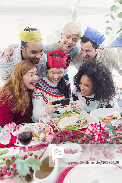 Freunde in Papierkronen  die beim Weihnachtsessen Selfie nehmen