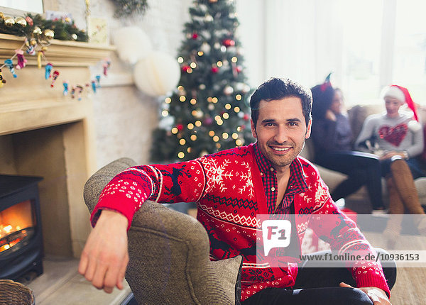 Portrait lächelnder Mann im Weihnachtspullover sitzend im Wohnzimmer