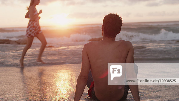 Junger Mann beobachtet Frau  die bei Sonnenuntergang in der Meeresbrandung läuft