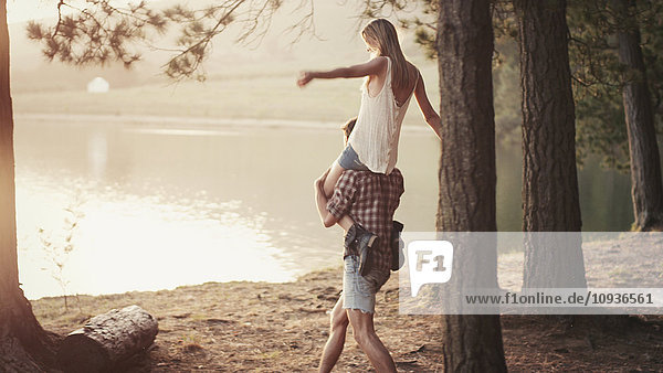 Junger Mann trägt Frau auf den Schultern am Seeufer