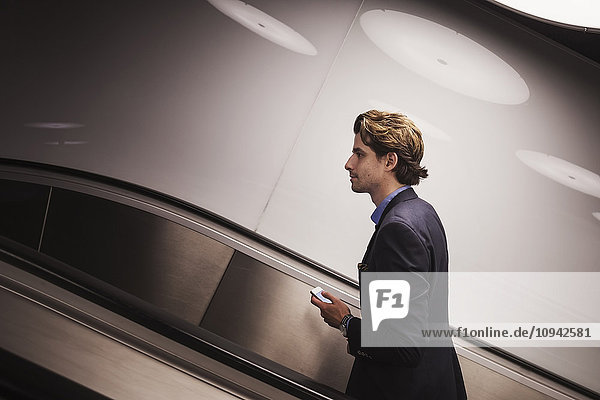 Seitenansicht des Geschäftsmannes mit Handy auf Rolltreppe am Flughafen