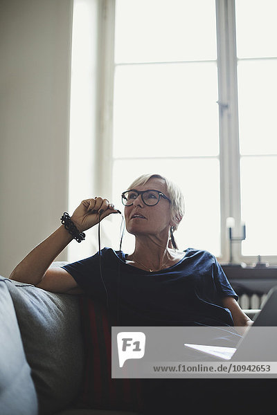 Architect holding in-ear headphones while sitting with laptop on sofa at home