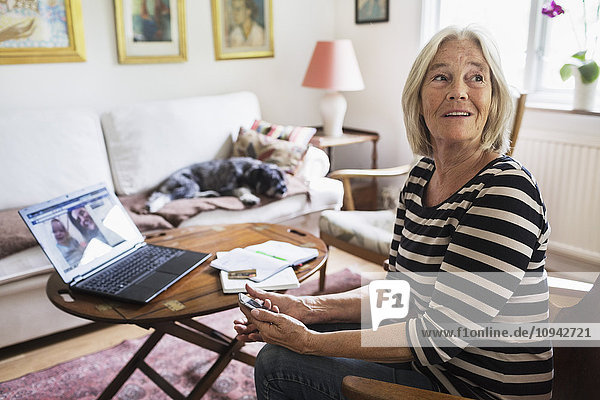 Happy senior woman looking away at home while dog relaxing on sofa