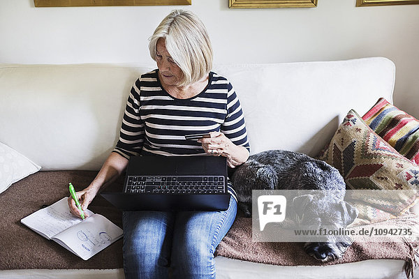 Seniorin schreibt im Buch  während sie Laptop und Kreditkarte von Hund auf dem Sofa zu Hause benutzt.