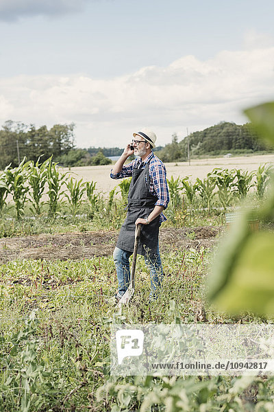 Gardener talking on mobile phone while standing with shovel at farm against sky