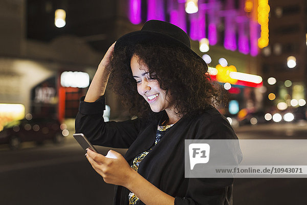 Lächelnde junge Frau liest Textnachrichten auf dem Smartphone in der Stadt bei Nacht.