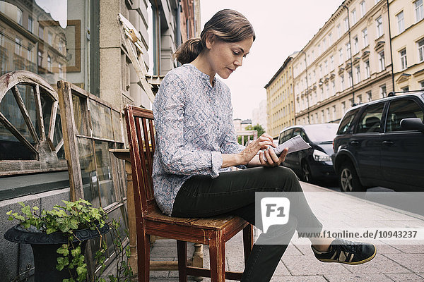 Frau liest Papier  während sie auf einem Stuhl gegen einen Laden in der Stadt sitzt.