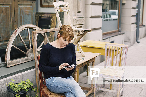 Frau  die das Handy benutzt  während sie auf dem Stuhl gegen den Laden sitzt.