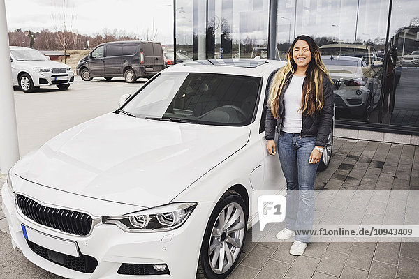 Portrait of smiling young woman standing by car against showroom