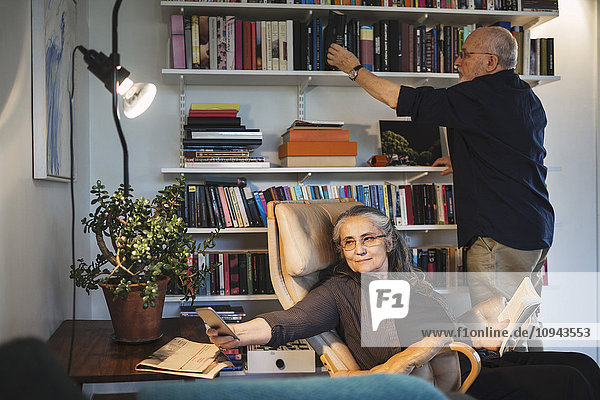 Seniorin nimmt Selfie  während der Mann Bücher im Regal arrangiert.