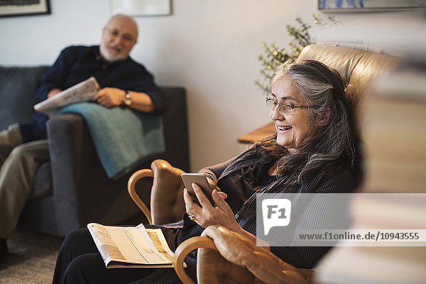 Glückliche Seniorin mit Handy  während der Mann zu Hause auf dem Sofa sitzt.