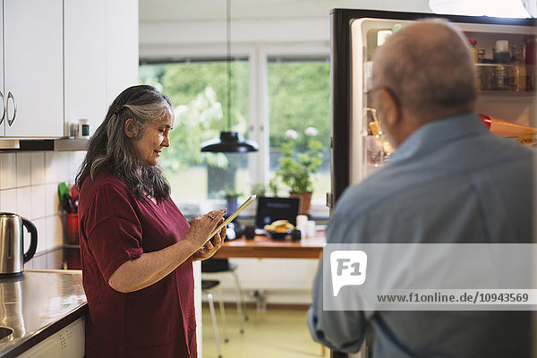 Side view of senior woman using digital tablet while man standing at refrigerator