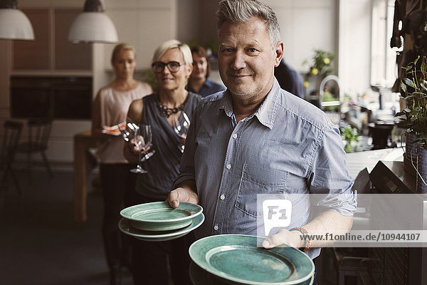 Portrait of mature man holding plates with friends in background at home
