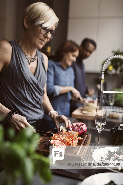 Reife Frau beim Obstschneiden mit Freunden im Hintergrund an der Küchentheke
