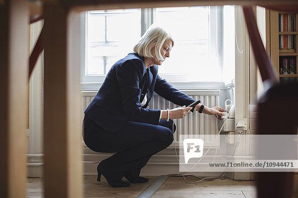 Senior woman crouching while charging mobile phone by radiator with furniture in foreground at library