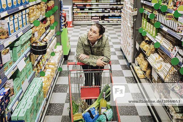 Hochwinkelansicht einer nachdenklichen Frau  die sich im Supermarkt auf den Einkaufswagen lehnt