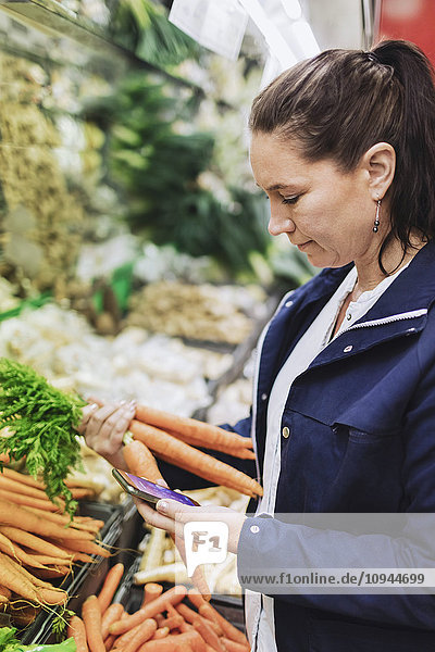 Seitenansicht der Frau mit Smartphone beim Kauf von Möhren im Supermarkt