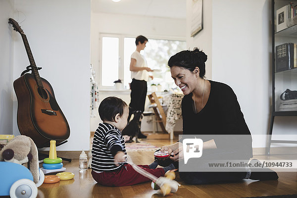 Mother with toddler in living room while woman working at kitchen
