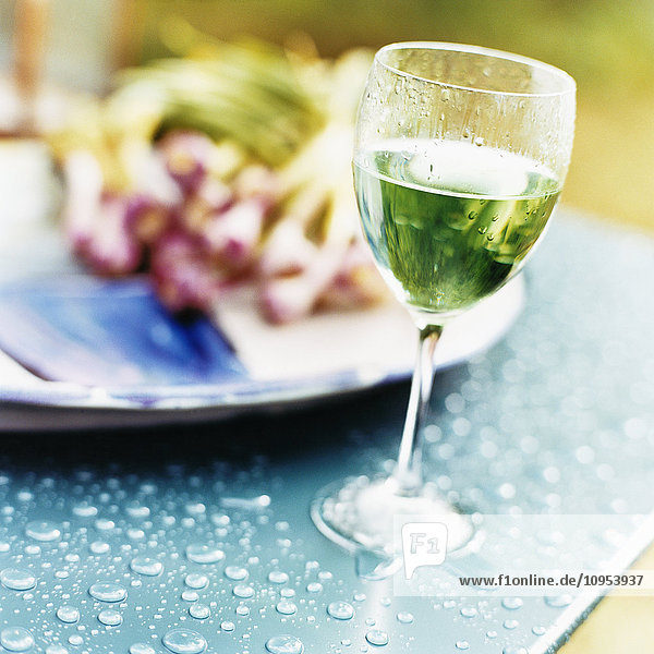 Nahaufnahme eines Weinglases mit Weißwein und einem Teller mit Speisen auf einem blauen Tuch.