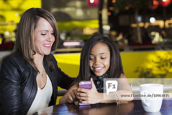 'Young mother and her daughter in a cafe looking at a smart phone while taking a break from shopping; St. Albert Alberta Canada'