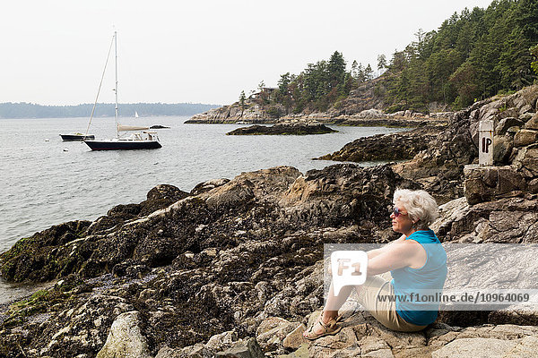 'Woman sitting on rocks at the water's edge looking out over the coast; Bowen Island  British Columbia  Canada'