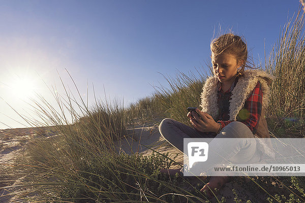 'A young girl sits on the sand of a beach holding a digital device; Tarifa  Cadiz  Andalusia  Spain'