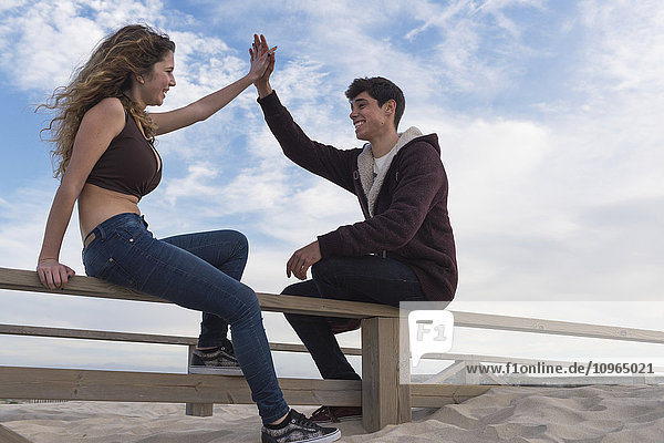 'A young man and woman sit talking and give a high five on a wooden fence at the beach; Tarifa  Cadiz  Andalusia  Spain'