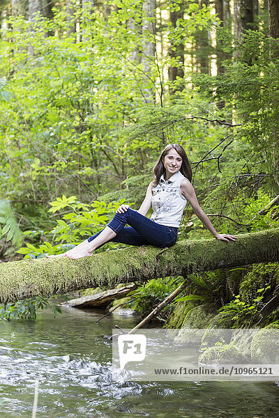 Porträt eines Mädchens im Teenageralter  das auf einem moosbewachsenen Baumstamm über einem Bach sitzt; Chilliwack  British Columbia  Kanada'.