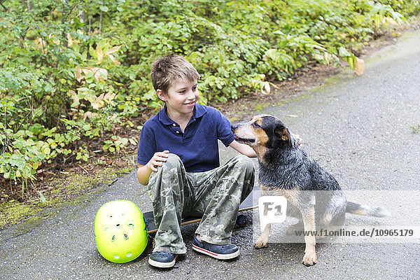 Junge  der auf einem Skateboard sitzt und seinen Hund streichelt; Salmon Arm  British Columbia  Kanada'.