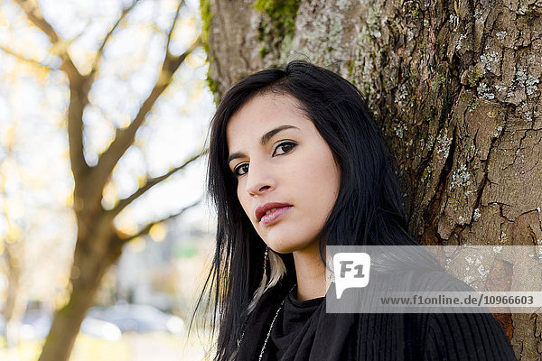 'A young aboriginal female model headshot in the outdoors in autumn with a tilted gaze into the camera leaning on a tree; Vancouver  British Columbia  Canada'