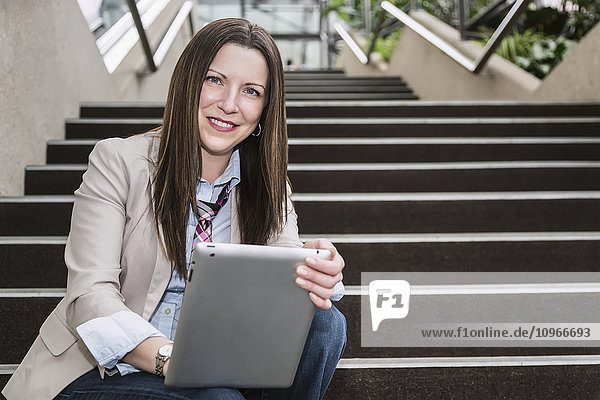 Porträt einer reifen Geschäftsfrau  die auf einer Treppe im Atrium eines Bürogebäudes mit einem Tablet sitzt; Edmonton  Alberta  Kanada'.