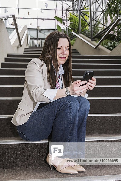 'Mature business woman sitting on stairway in the atrium of an office building and texting on her smart phone; Edmonton  Alberta  Canada'