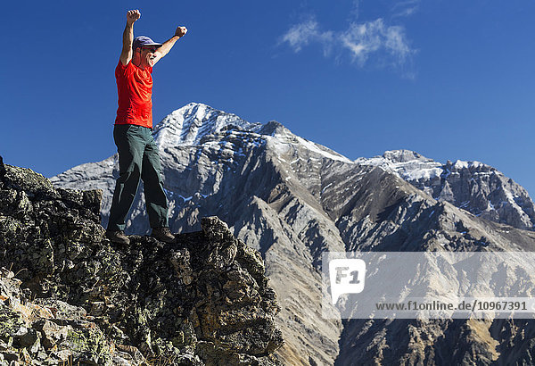 'Male hiker standing with arms raised in the air on top of mountain ridge overlooking snow peaked mountains with blue sky and clouds  Kananaskis Provincial Park; Alberta  Canada'