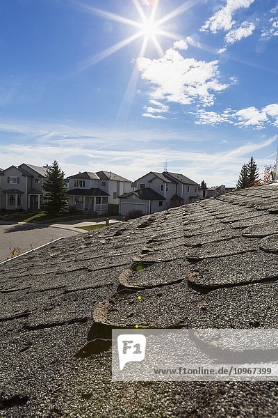 'Close up of well worn rounded roof shingles with neighbourhood houses sun burst blue sky and clouds in the background; Calgary Alberta Canada'
