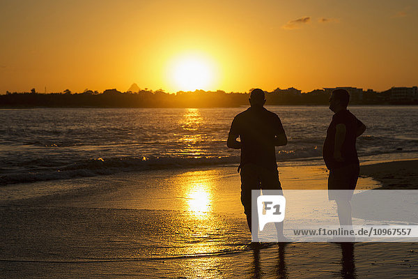 'Two men stand in the tide water on a beach at sunset; Caloundra  Queensland  Australia'