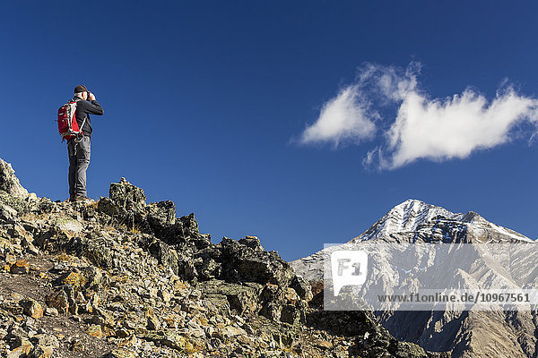 'Male hiker standing on top of mountain ridge looking through binoculars at distant snow peaked mountain with blue sky and clouds  Kananaskis Provincial Park; Alberta  Canada'