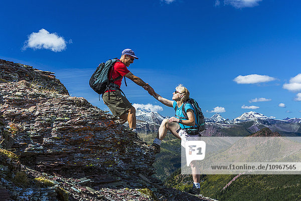 'Male hiker helping female hiker up a rocky peak of a mountain ridge with grand vista and blue sky and clouds in the background; Waterton  Alberta  Canada'