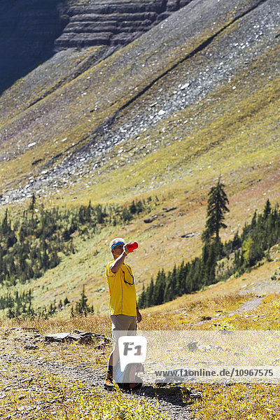 'Male hiker on trail taking a drink of water from water bottle with sloping mountain side in the background; Waterton  Alberta  Canada'