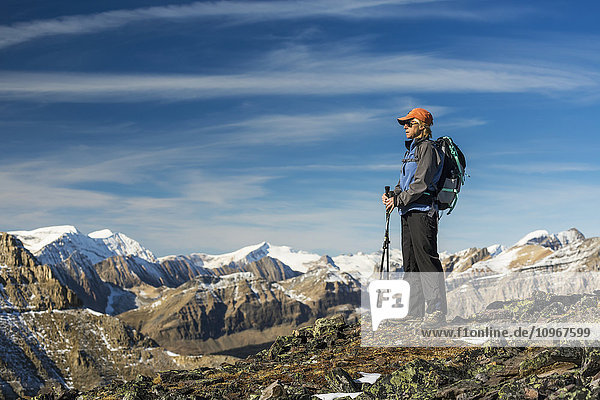 'Female hiker with poles standing on top of a rocky mountain ridge overlooking snow peaked mountain ranges with blue sky and clouds  Banff National Park; Alberta  Canada'